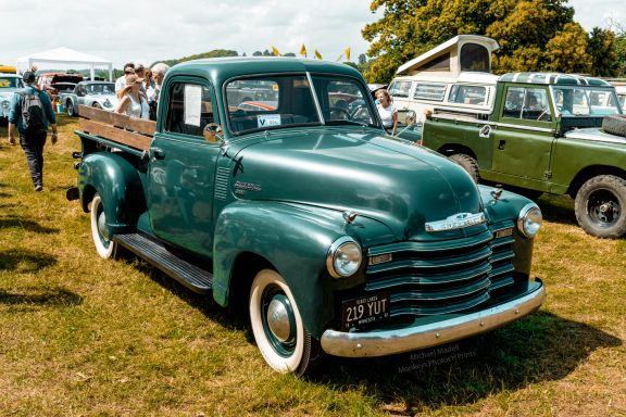 Classic green pickup truck parked on grass, surrounded by vintage vehicles and people.