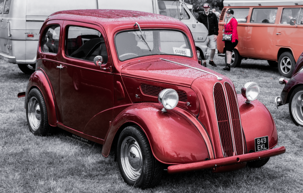 Red vintage car parked on grass with blurred background vehicles.