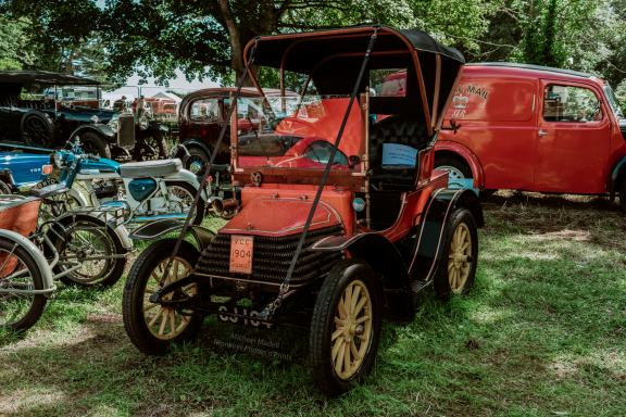 A vintage red car with yellow wheels, surrounded by classic vehicles in a grassy area.